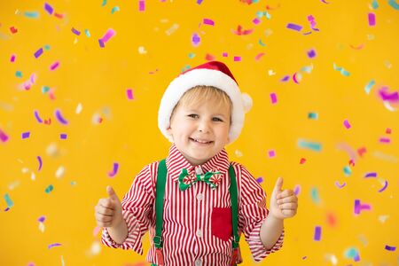 Portrait of happy child showing thumbs up. Funny kid dressed Santa Claus hat against yellow background. Christmas holiday conceptの写真素材