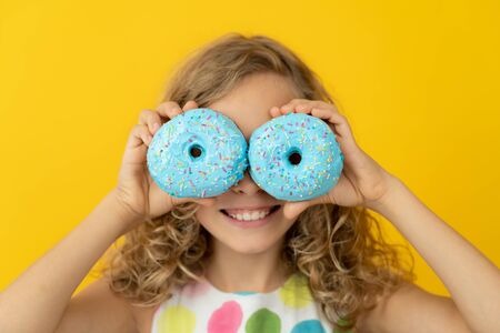 Happy child holding glazed donut. Portrait of funny girl against yellow backgroundの写真素材