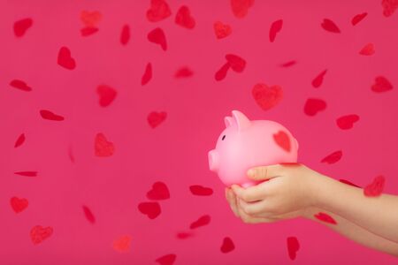 Piggy bank in children hands with red heart shaped confetti against pink background. Valentineâs day conceptの写真素材