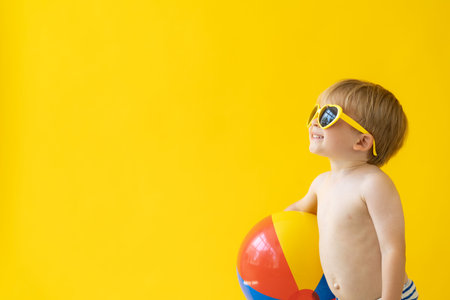 Portrait of happy child holding beach ball against yellow background. Kid having fun on summer...