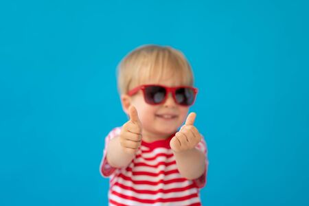Close up portrait of happy child wearing striped shirt against blue background. Kid having fun on summer vacation. Boy showing thumbs upの写真素材