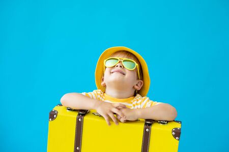 Portrait of happy child with yellow suitcase against blue background. Kid having fun on summer vacation. Dream and imagination conceptの写真素材
