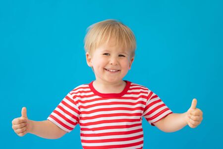 Close up portrait of happy child wearing striped shirt against blue background. Kid having fun on summer vacation. Boy showing thumbs upの写真素材