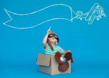 Happy child dreams of becoming a pilot. Kid having fun against blue paper background. Boy wearing striped shirt playing in cardboard box. Summer vacation and travel concept. Dream and imaginationの写真素材