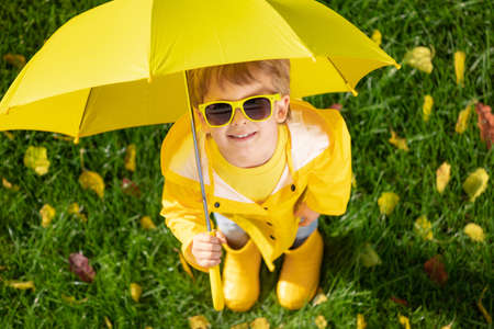 Top view portrait of happy child with umbrella outdoor in autumn park. Smiling kid standing on green grass against yellow leaves blurred backgroundの写真素材
