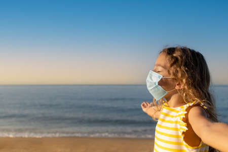Happy child wearing medical mask outdoor against blue sky background. Girl enjoying by sea at summer. Coronavirus pandemic and healthy lifestyle conceptの写真素材