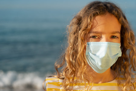 Serious child wearing medical mask outdoor against blue sky background. Girl enjoying by sea at...