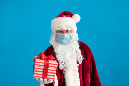 Senior wearing Santa Claus costume and protective mask. Man holding gift box. Christmas holiday...