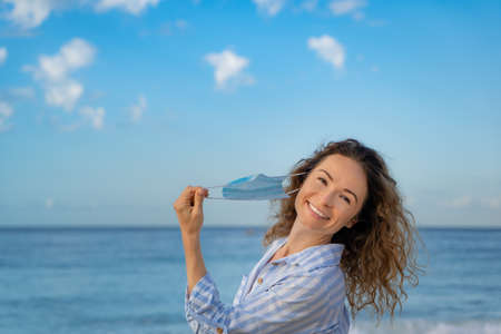 Happy woman wearing medical mask outdoor against blue sky background. Person enjoying by sea at summer. Coronavirus pandemic and healthy lifestyle conceptの写真素材