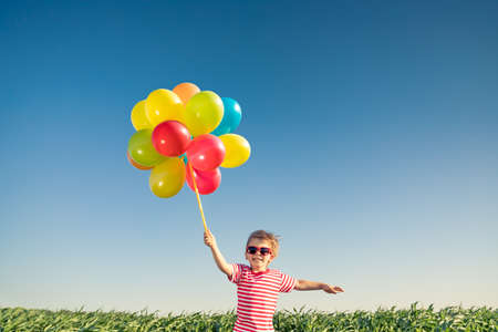 Happy child playing with bright multicolor balloons outdoor. Kid having fun in green spring field against blue sky background. Healthy and active lifestyle conceptの写真素材