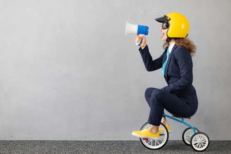 Successfull businesswoman driving toy car outdoor. Funny young woman shouting through megaphone against concrete wall background. Business srart up and winner conceptの写真素材