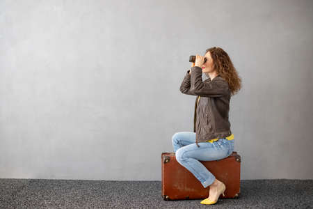 Young woman is ready to travel. Funny woman sitting on retro suitcase against concrete wall background. Summer vacation during pandemic coronavirus conceptの写真素材