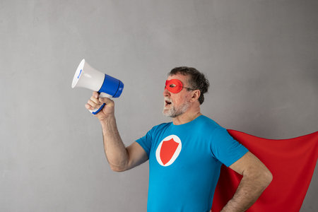 Senior wearing superhero costume against grey concrete wall background. Man shouting through...