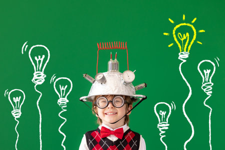 Smart child against green chalkboard. Funny kid wearing helmet with lightbulb. Education, start up and business idea conceptの写真素材
