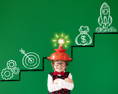 Smart child against green chalkboard. Funny kid wearing helmet with lightbulb. Education, start up and business idea conceptの写真素材