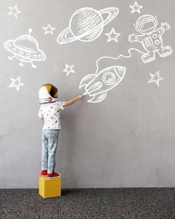 Dream big! Happy child playing outdoor. Funny kid draws a chalk rocket on the wall. Child dreaming about space. Kid pretend to be astronaut. Children dream and imagination conceptの写真素材