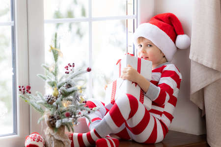 Child sitting on windowsill. Happy kid wearing Christmas pajamas. Child holding xmas gift. Portrait of smiling kidの写真素材