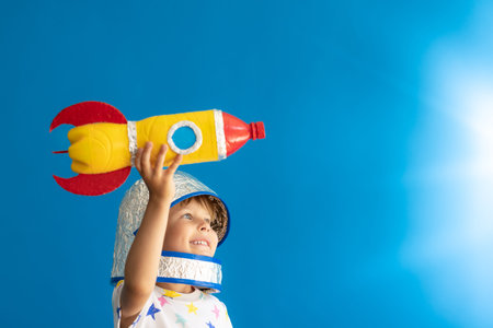 Happy child playing with toy rocket against blue background. Kid pretend to be astronaut....