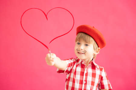 Happy child painting red heart. Portrait of smiling kid against pink background. Valentines day holiday conceptの写真素材