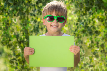 Happy child showing paper blank. Portrait of kid outdoor. Boy against blurred green spring background. Earth day and ecology conceptの写真素材