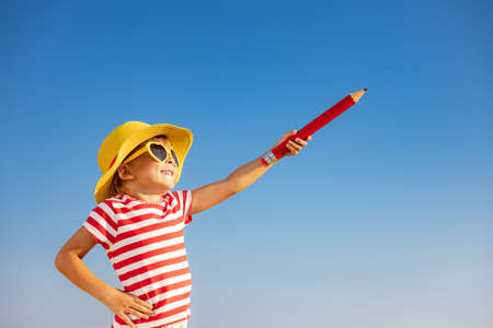 Happy child holding giant pencil outdoor against blue sky background. Portrait of smiling kid on summer vacation. Freedom and travel conceptの写真素材