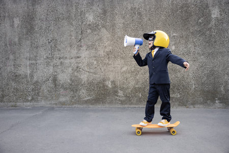 Happy child pretend to be businessman. Funny kid riding skateboard outdoor. Boy shouting through...