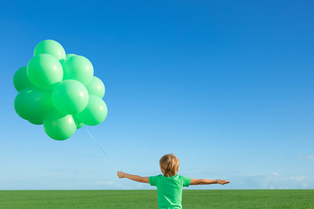 Happy child playing with bright multicolor balloons outdoor. Kid having fun in green spring field against blue sky background. Healthy and active lifestyle conceptの写真素材