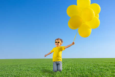Happy child jumping in green spring field. Kid with yellow balloons against blue sky background. Carefree and freedom outdoorの写真素材