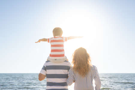 Happy family having fun on the beach. Mother, father and child against blue sea and sky background. Summer vacation concept. Rear view portraitの写真素材