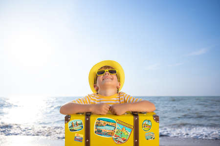 Happy child enjoying the sun against blue sky background. Kid with suitcase having fun on the beach. Summer vacation and holiday conceptの写真素材
