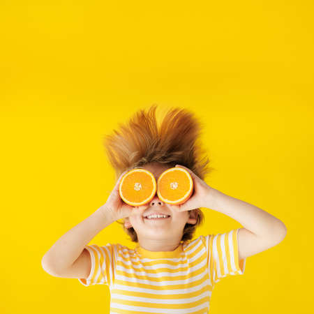 Surprized child holding slices of orange fruit like sunglasses. Happy kid wearing striped yellow t-shirt against paper background. Healthy eating and summer vacation conceptの写真素材