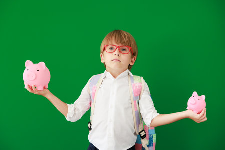 Funny child student holding piggy bank in class. Happy kid against green chalkboard. Education and back to school conceptの写真素材