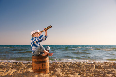 child pretend to be sailor. Kid looking through spyglass. Child sitting on old barrel on the beach. Boy on summer vacation. adventure and travel conceptの写真素材