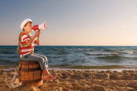 Child pretend to be a sailor. Kid holding loudspeaker. Child sitting on old barrel on the beach. Boy on summer vacation. Adventure and travel conceptの写真素材