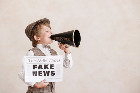 Newsboy shouting through loudspeaker against grunge wall background. Boy selling fake news. Child wearing vintage costume. Kid holding newspaper. Social media and Internet nerve work conceptの写真素材