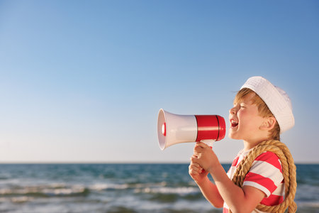 Happy child pretend to be sailor. Kid agains blue sea and sky. Boy on summer vacation. Child shouting through loudspeaker. Adventure and travel conceptの写真素材