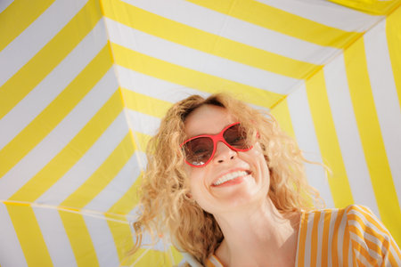 Happy young woman under a striped yellow parasol. Low angle view portrait of a girl laughing at the camera. Female wearing sunglasses enjoys a summer vacationの写真素材