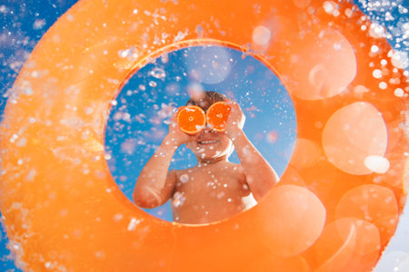 Happy child holds the orange halves like sunglasses. Kid having fun on summer vacation. Boy jumping...