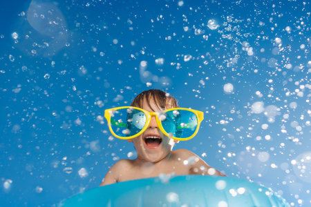 Happy child playing in waterpark. Kid having fun on summer vacation. Boy jumping into swimming pool with splashes of water. Funny child wearing sunglasses against blue sky backgroundの写真素材