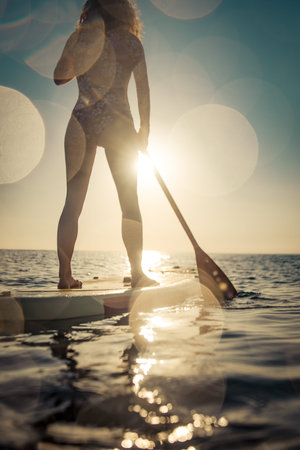 Silhouette of woman walking on stand-up paddleboard by the sea. Female enjoying by sunrise during...