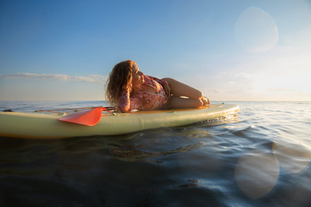 Happy woman walking on stand-up paddleboard by the sea. Female enjoying by sunrise during summer...