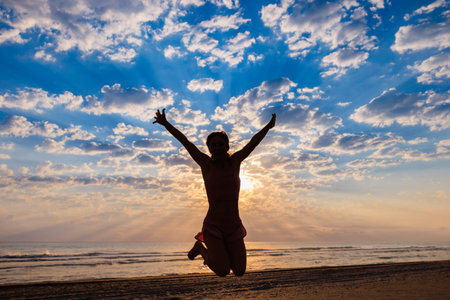 Fit happy woman enjoying the sunrise at the beach. Young female jumping against sea and sky...