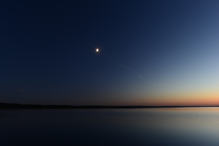Moon in the dark night sky moonlight with the afterglow of the twilight sunset over the quiet surface of the lake waterの写真素材