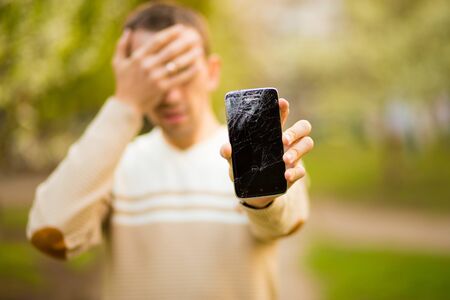 Handsome young man hold broken smartphone screen stressed with hand on head, shocked with shame and surprise face, angry and frustrated. Fear and upset for mistake.の写真素材