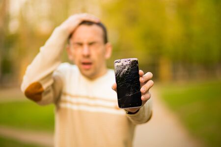 Handsome young man hold broken smartphone screen stressed with hand on head, shocked with shame and surprise face, angry and frustrated. Fear and upset for mistake.の写真素材