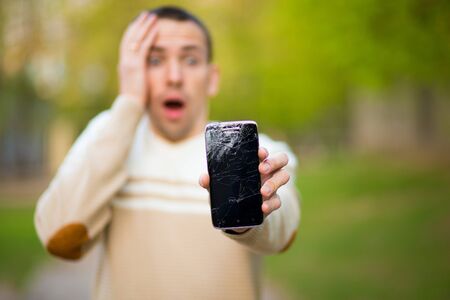 Handsome young man hold broken smartphone screen stressed with hand on head, shocked with shame and surprise face, angry and frustrated. Fear and upset for mistake.の写真素材