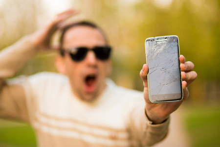 Handsome young man hold broken smartphone screen stressed with hand on head, shocked with shame and surprise face, angry and frustrated. Fear and upset for mistake.の写真素材