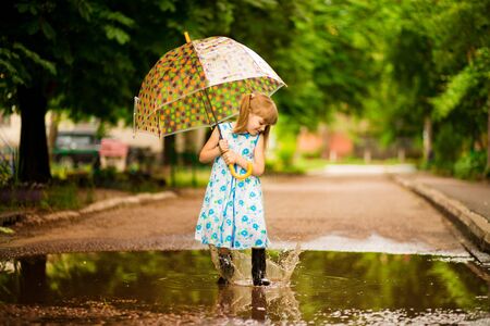 Happy funny kid girl with umbrella jumping on puddles in rubber boots and in polka dot dress and laughingの写真素材