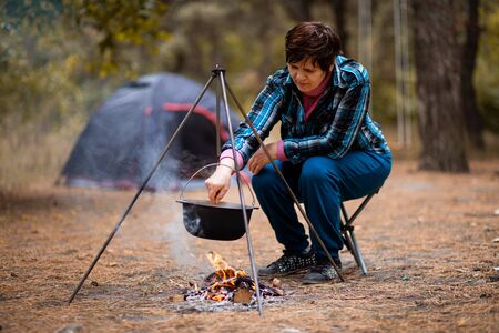 Senior woman preparing food on campfire while living in tent life in forest.の写真素材