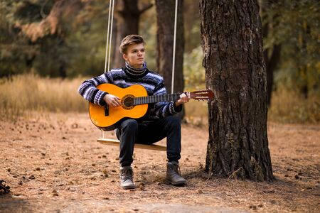 young handsome attractive man in sweater playing guitar in forest sitting on swing. Casual man, life style. Beautiful view on forestの写真素材